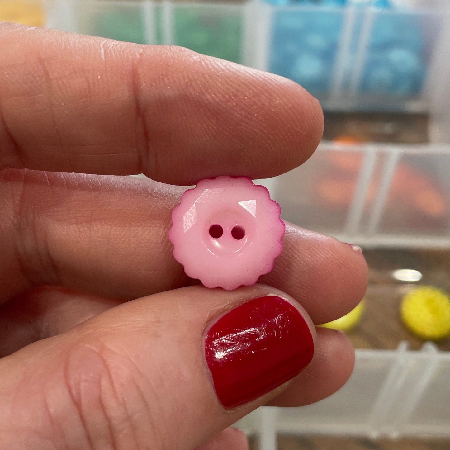 Hand holding a pink button with two holes in front of a container of buttons.