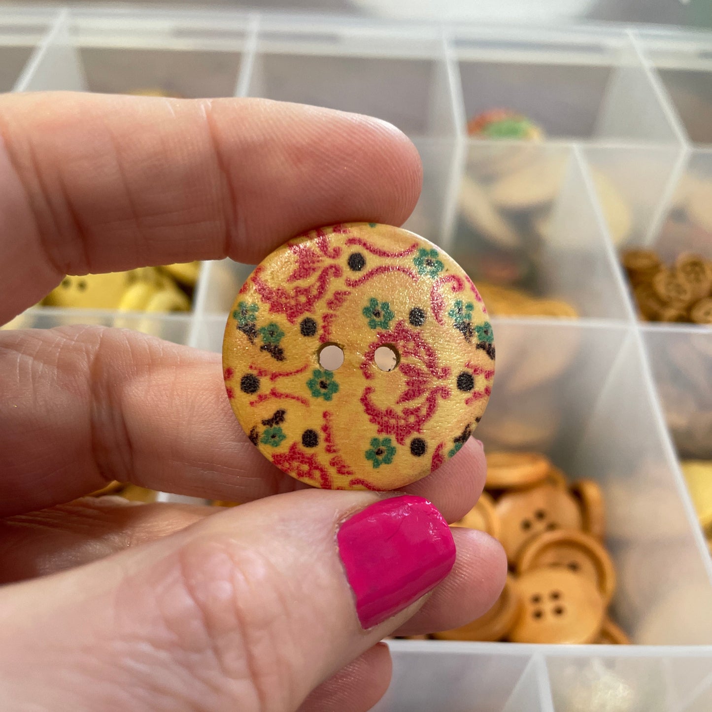 Hand holding a decorative button with a colorful pattern in front of a container of buttons.