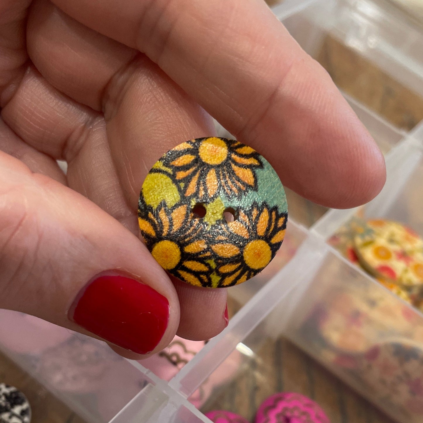 Hand holding a button with floral design in front of a container of assorted buttons.