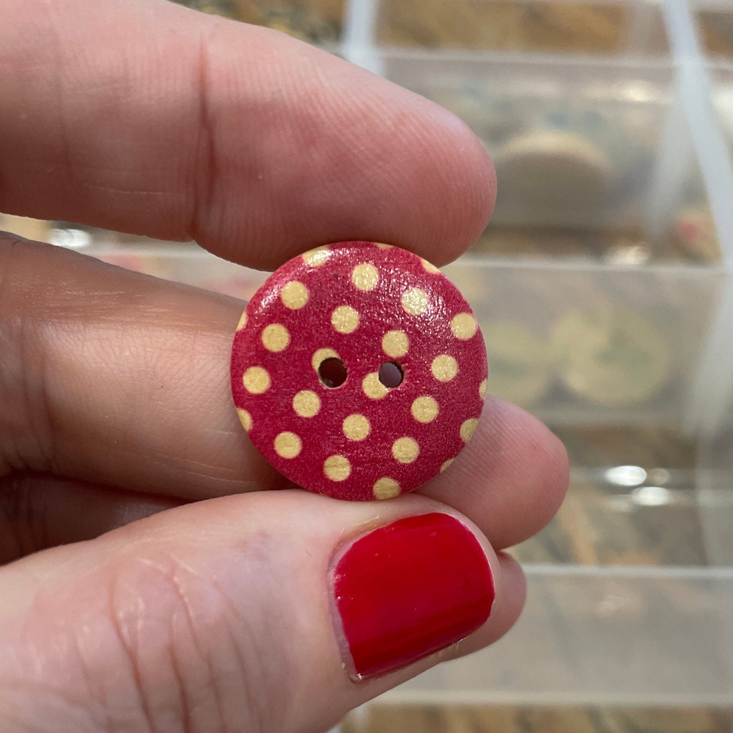Hand holding a red button with yellow polka dots in front of a clear plastic organizer.