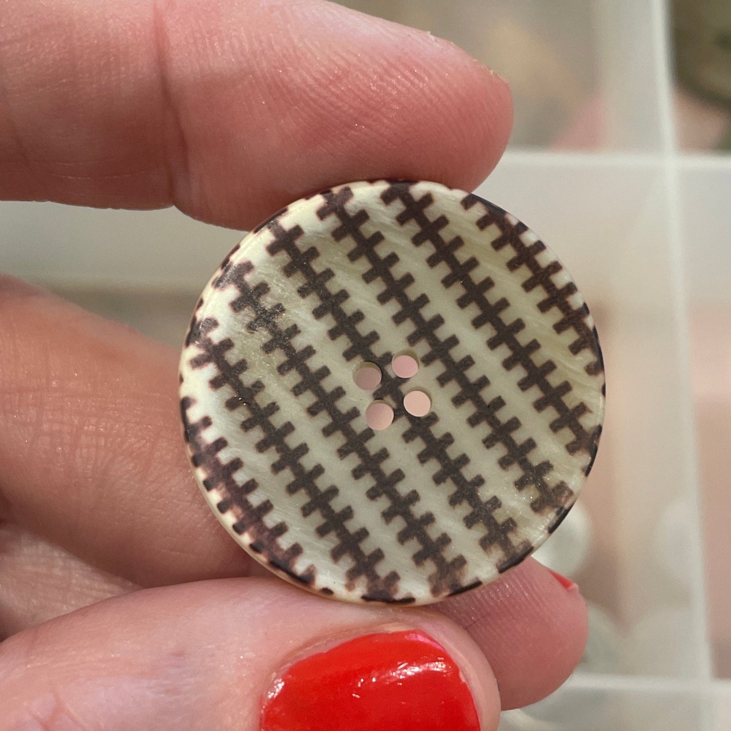 Hand holding a round button with a checkered pattern in front of a container of buttons.