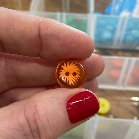 Hand holding a small orange object with a design, against a background of craft supplies.