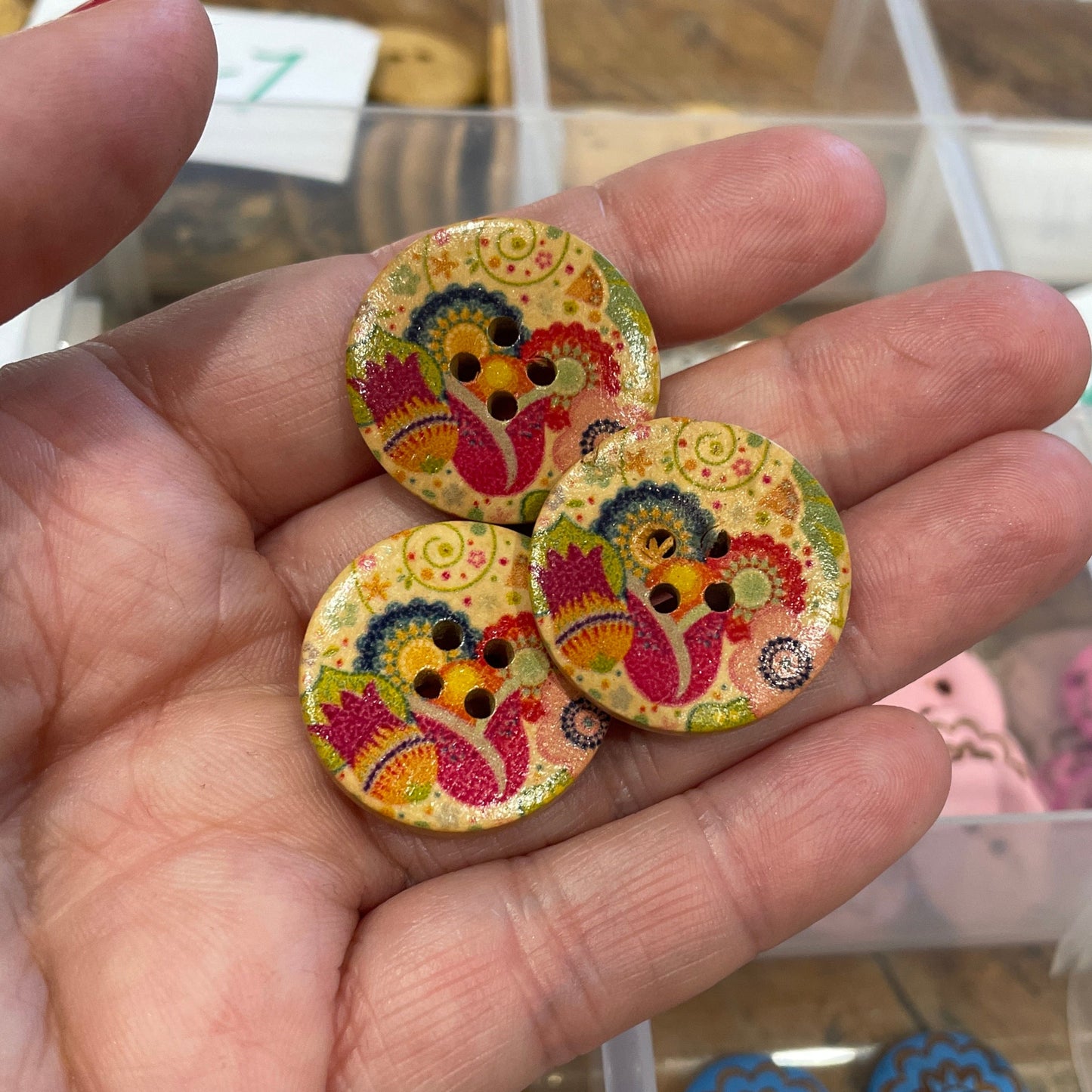Colorful decorative buttons held in a hand with a blurred background of a storage box.