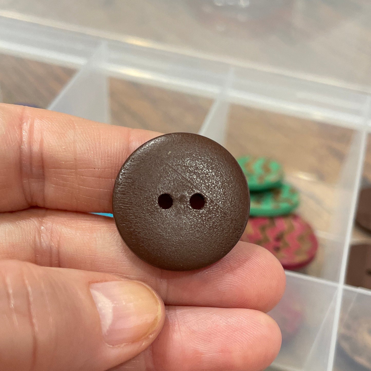 Hand holding a brown button with a container of various buttons in the background