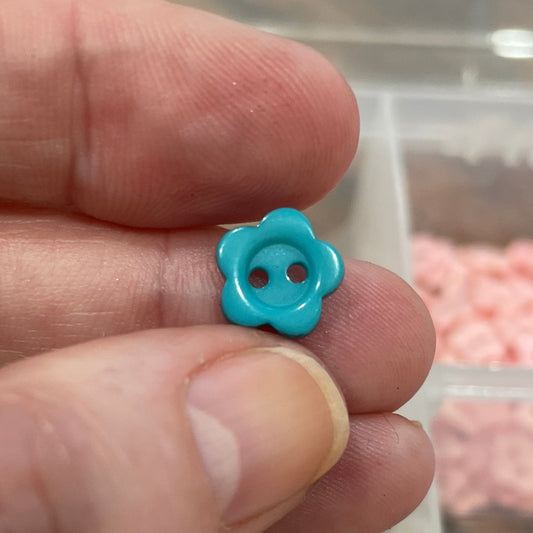 Hand holding a small blue flower-shaped button with a blurred background of colorful buttons.