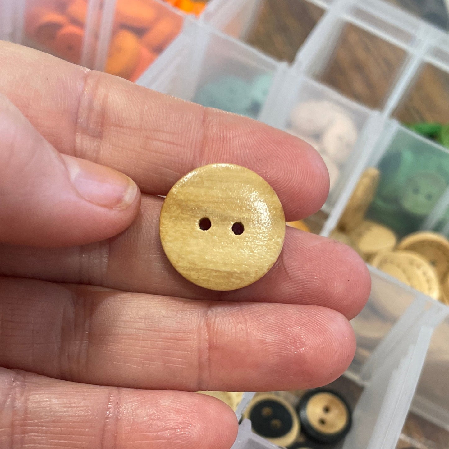 Hand holding a wooden button with a container of assorted buttons in the background