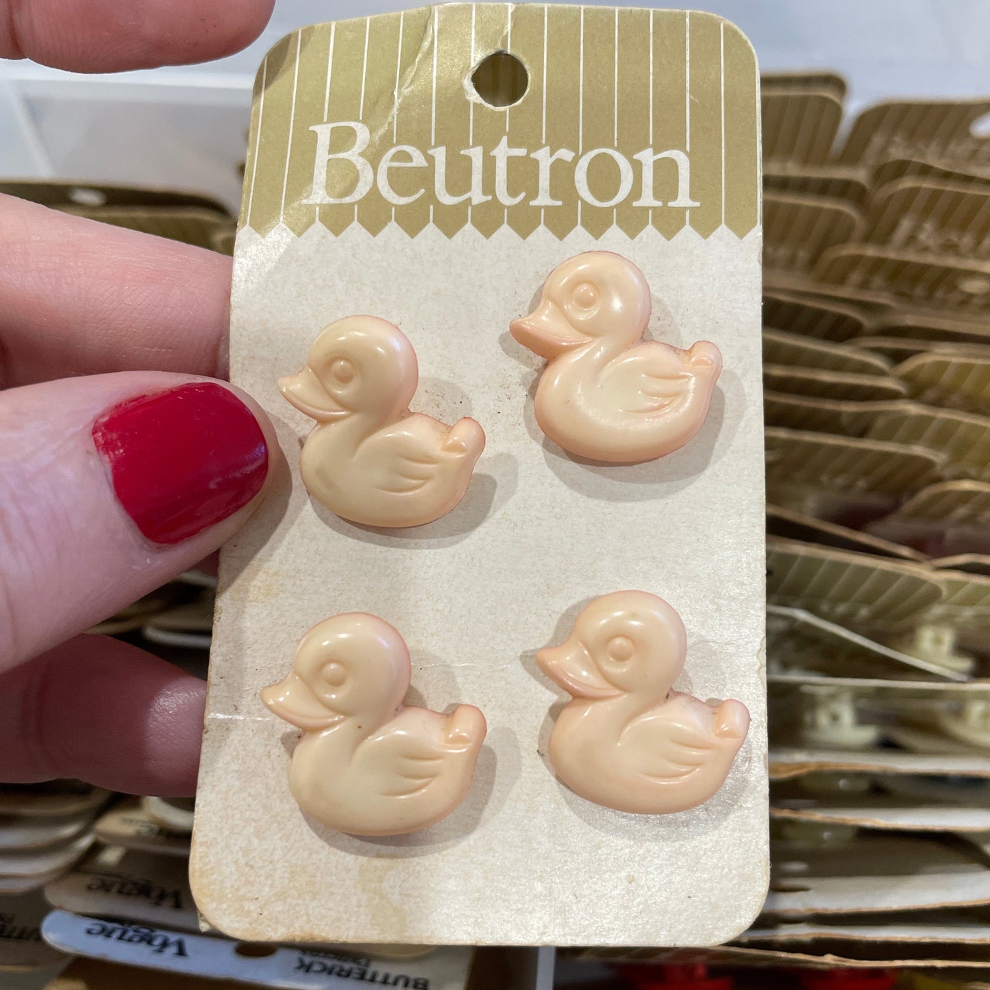 Set of small duck-shaped buttons on a Beutron branded card held by a hand with red nail polish.