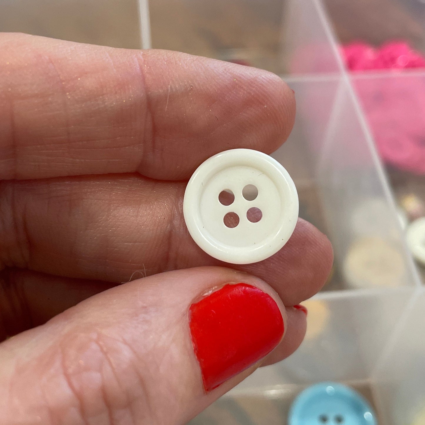 Hand holding a white button with four holes in front of a container of colorful buttons.