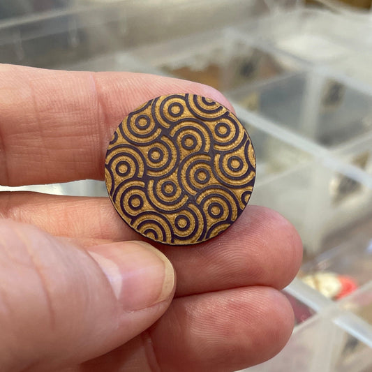 Hand holding a round wooden button with a pattern, against a blurred background of a container with similar buttons.