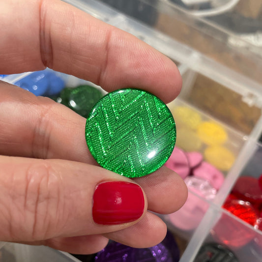 Hand holding a green textured button with a container of various buttons in the background