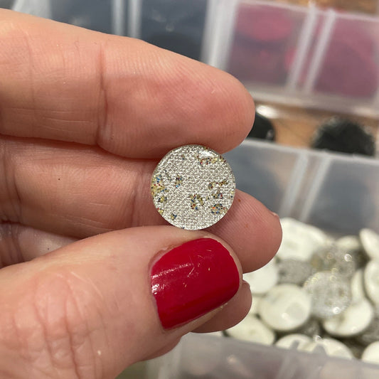 Hand holding a silver earring with a clear container of colorful buttons in the background