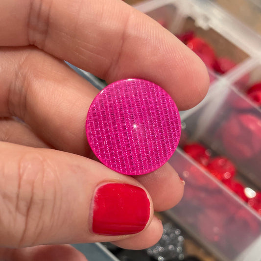 Hand holding a pink textured button with a container of various buttons in the background
