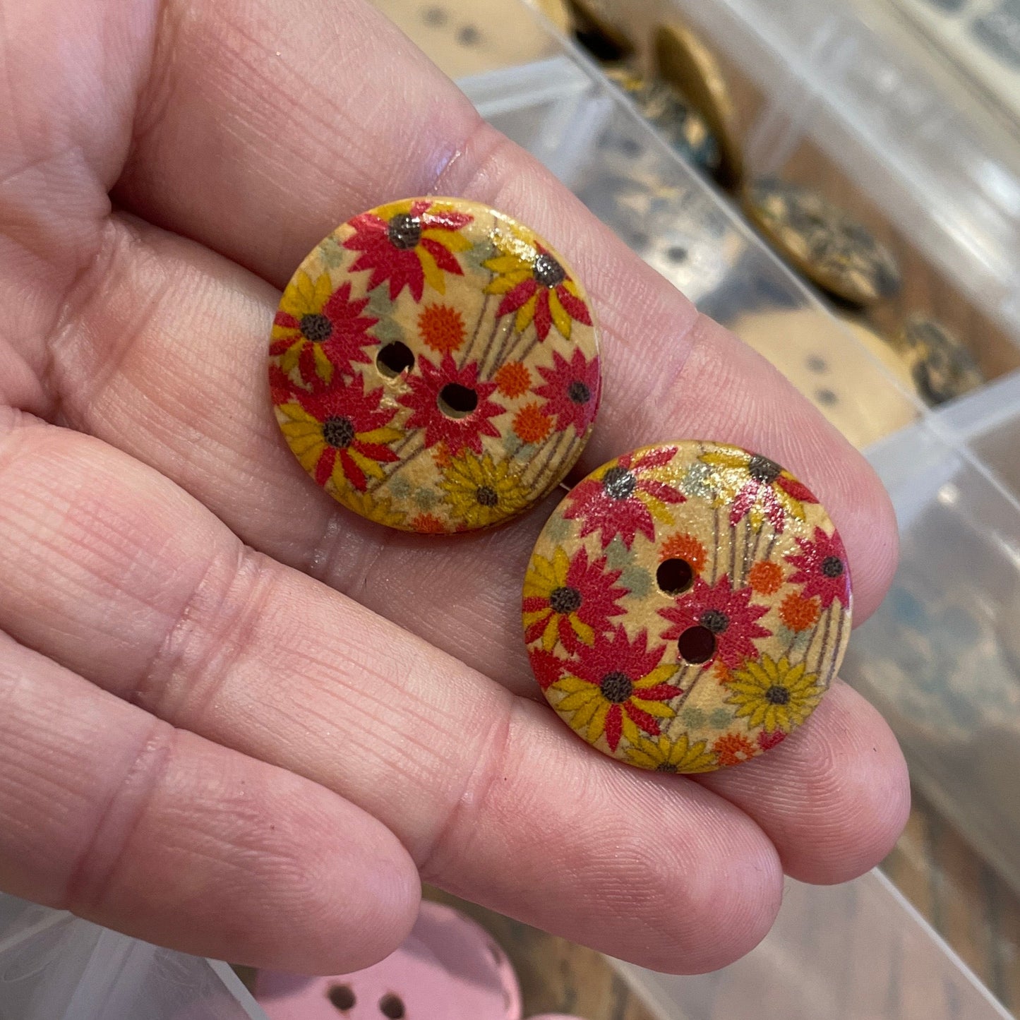 Hand holding two floral-patterned buttons with a container of assorted buttons in the background.