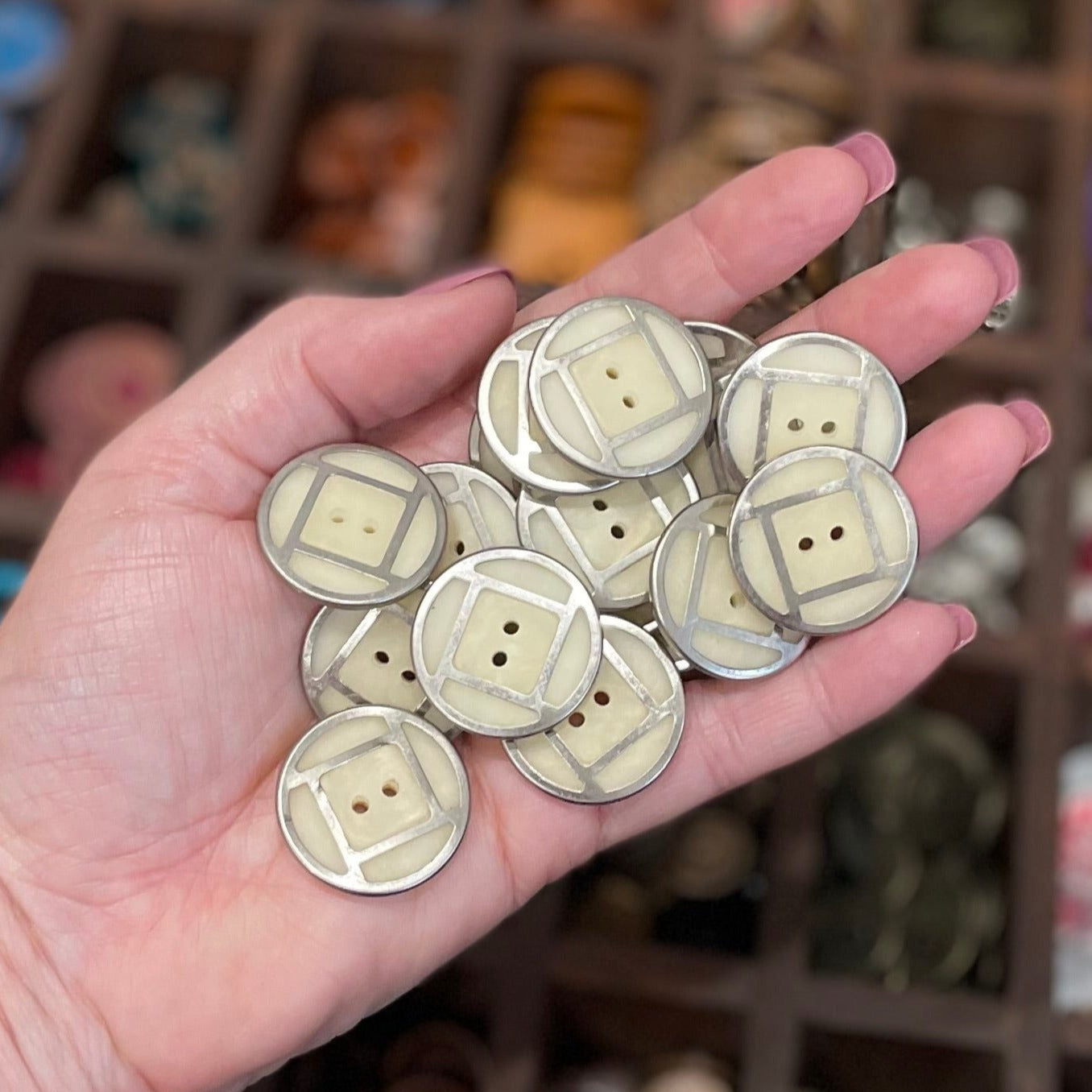 A hand holding several vintage cream-colored metal acrylic buttons with a blurred background of various trinkets.