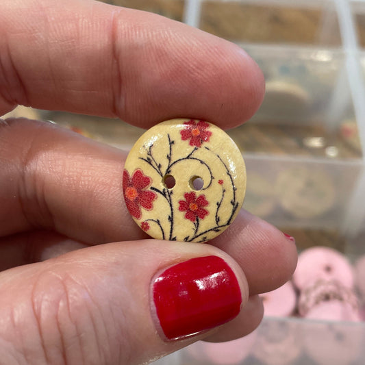 Hand holding a decorative button with a floral design in front of a container of buttons.