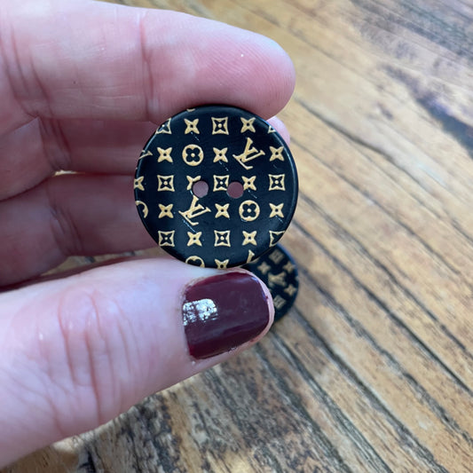 Black button with gold pattern held between fingers on a wooden surface