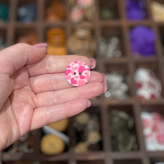 A hand holding a few pale pink resin buttons with a heart pattern and two holes each.