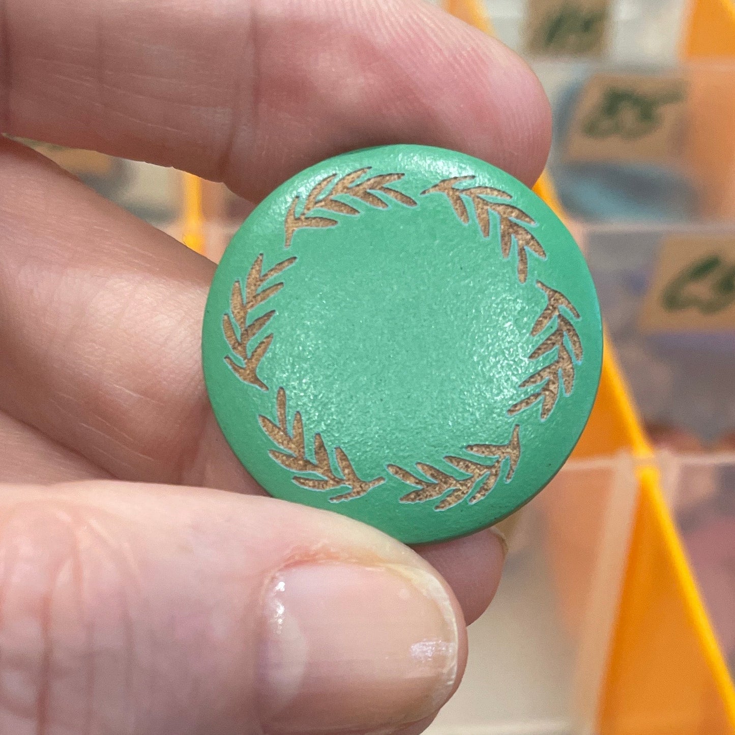 Green decorative button held by a hand with a blurred background of colorful buttons in a case.
