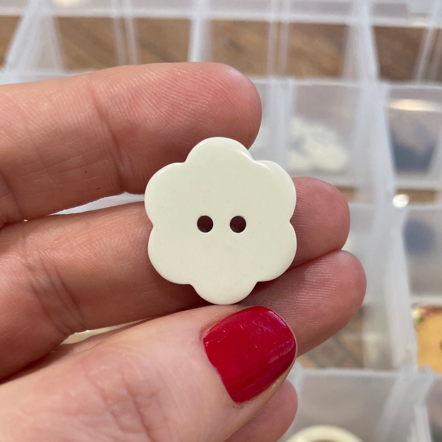 Hand holding a white floral button with a blurred container of buttons in the background