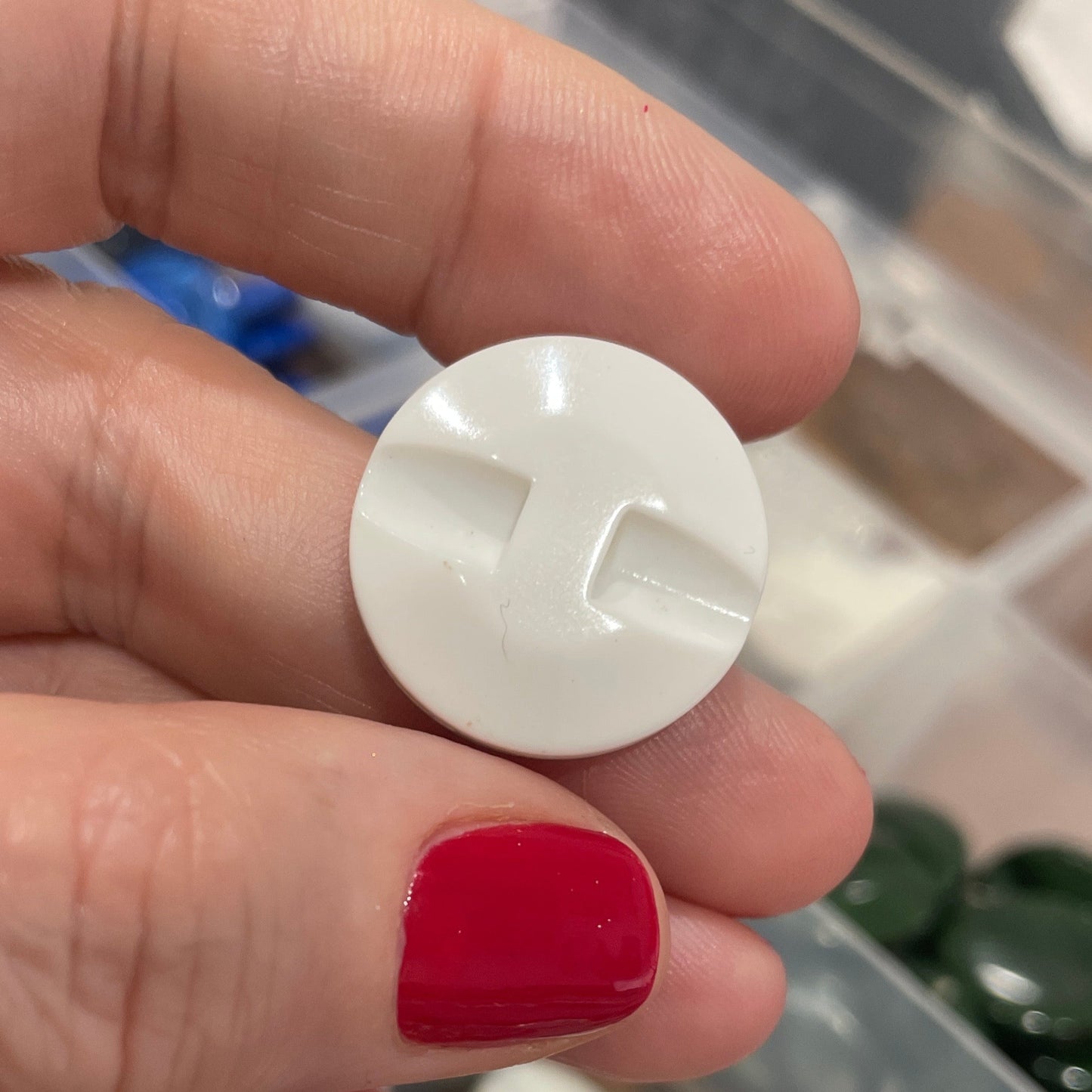 Hand holding a white circular object with a cross symbol, red nail polish visible, blurred background of a container with various items.