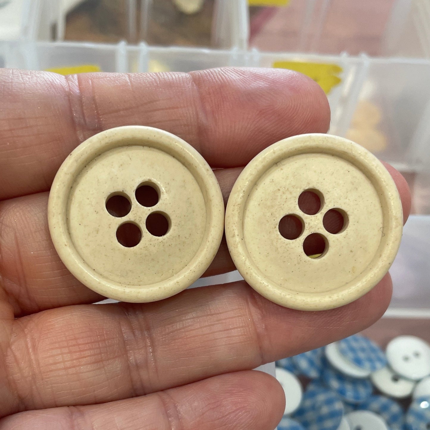 Two beige buttons held between fingers with a background of button containers.