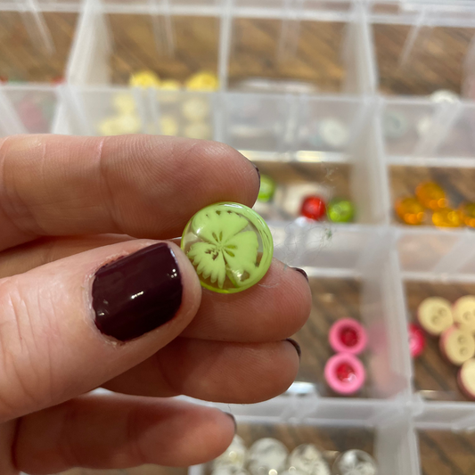Hand holding a small green button with a textured design, against a background of a clear plastic container with various buttons.