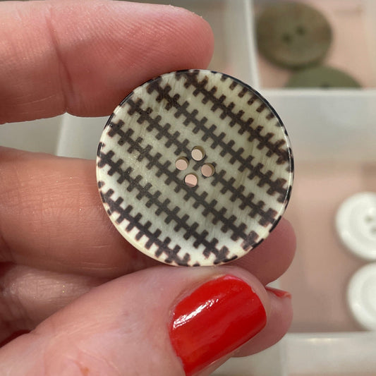 Hand holding a round button with a checkered pattern in front of a container of buttons.