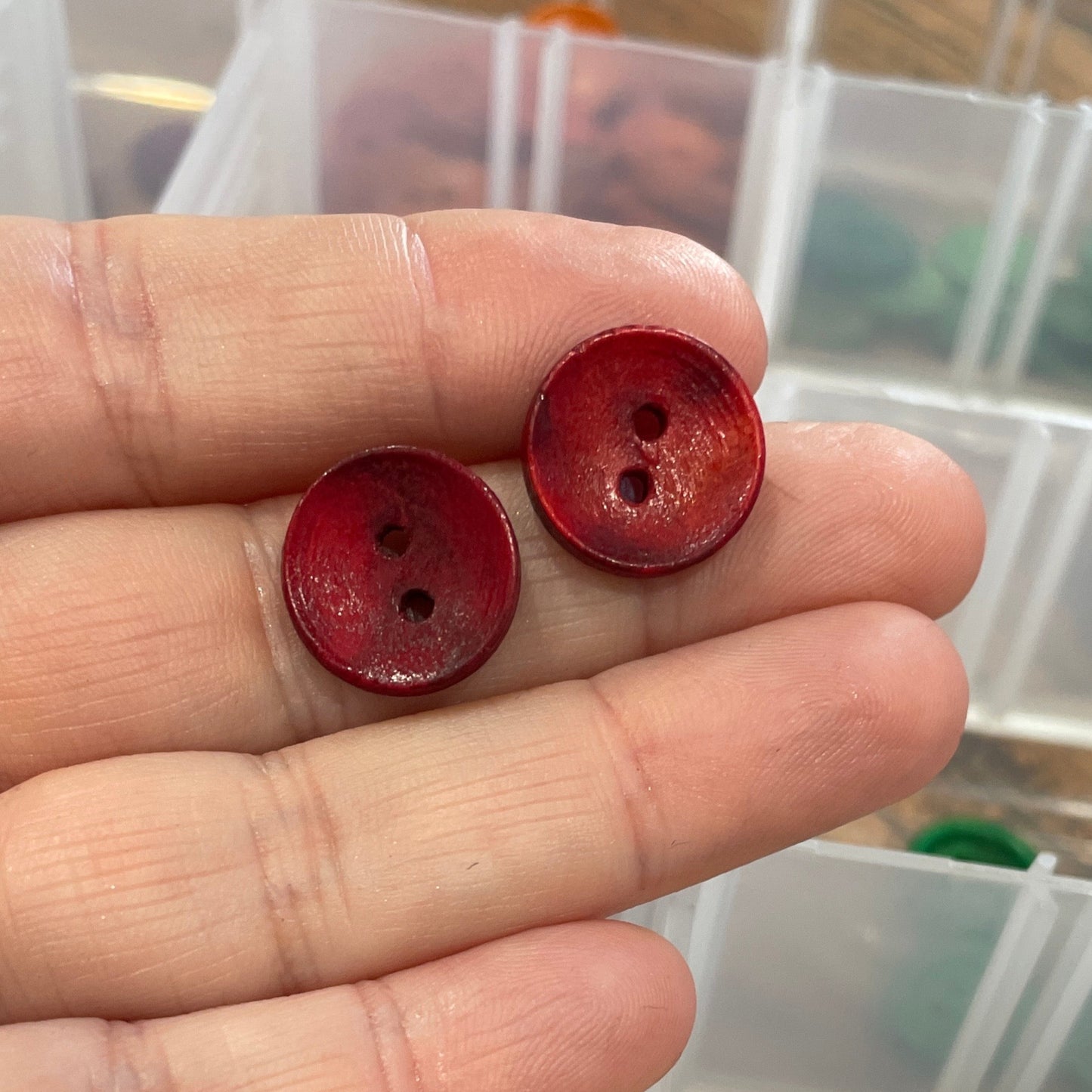 Hand holding two red buttons with a container of assorted buttons in the background.