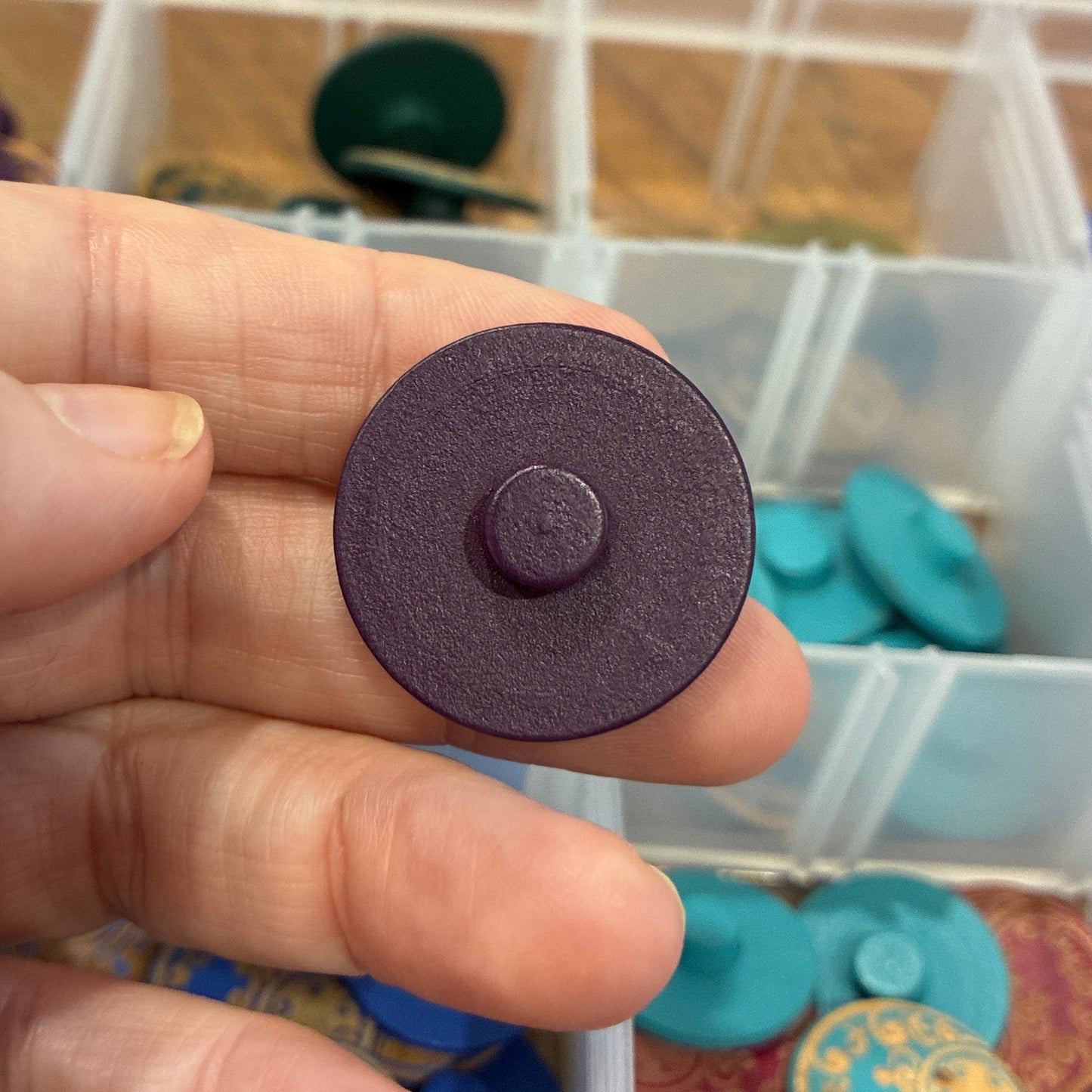 Hand holding a round purple button with a background of button containers.