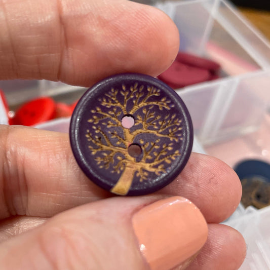 Hand holding a decorative button with intricate design, with a container of buttons in the background.