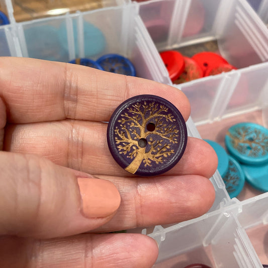 Hand holding a decorative button with a tree design in front of a container of assorted buttons.