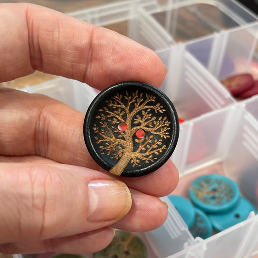 Hand holding a button with a tree design over a container of various buttons.