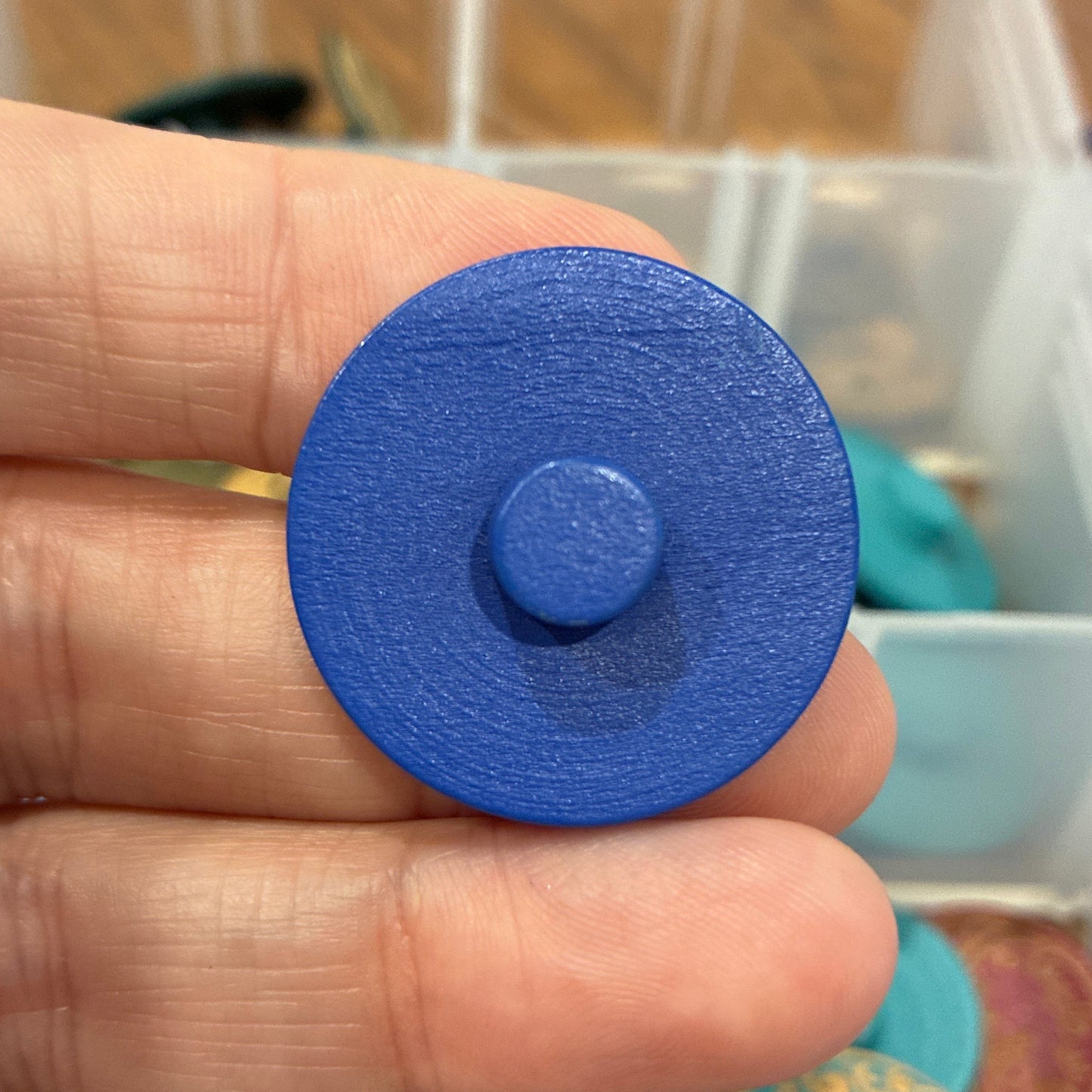Blue circular button held between fingers with a container of assorted colorful objects in the background