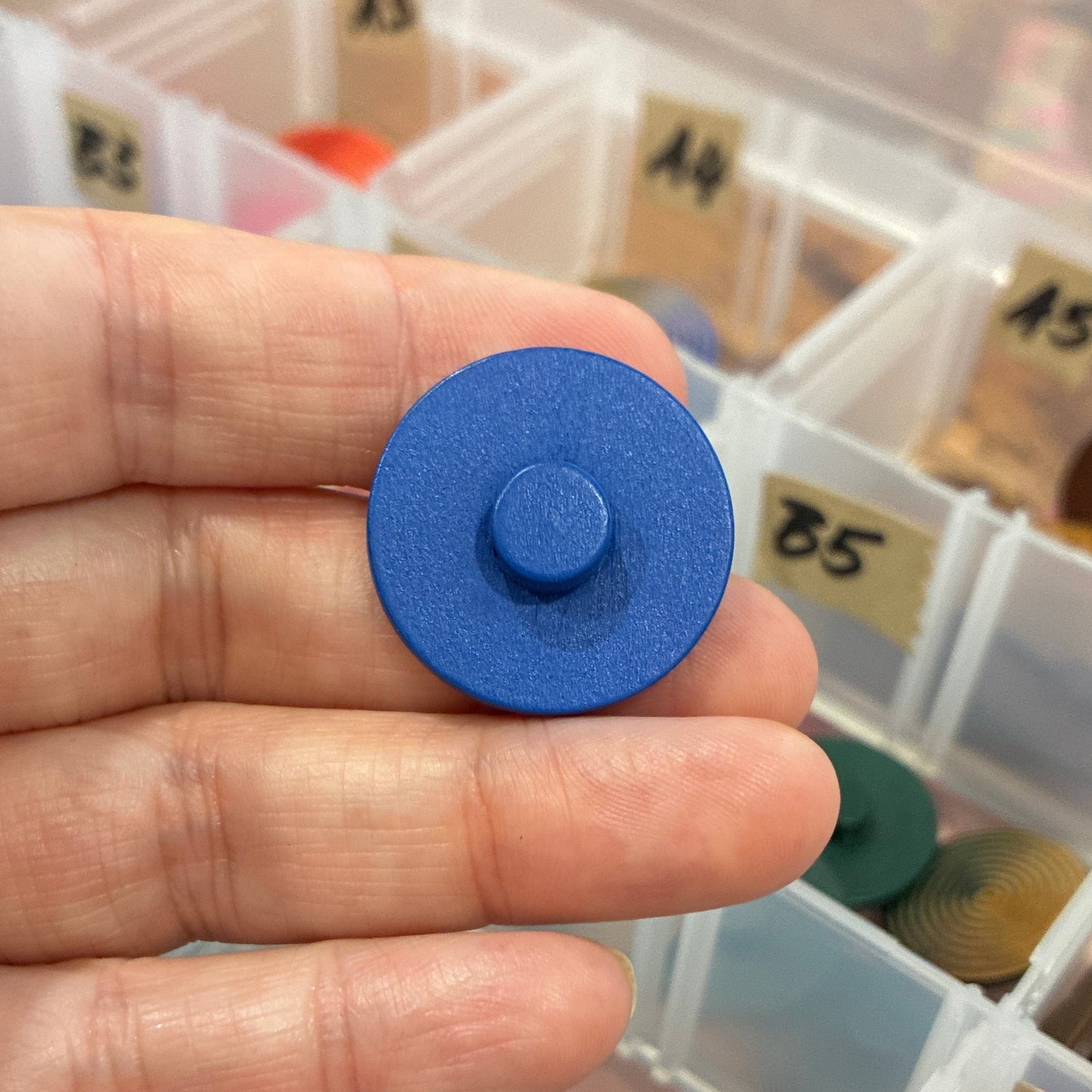 Blue circular button held in a hand with a container of assorted colored objects in the background