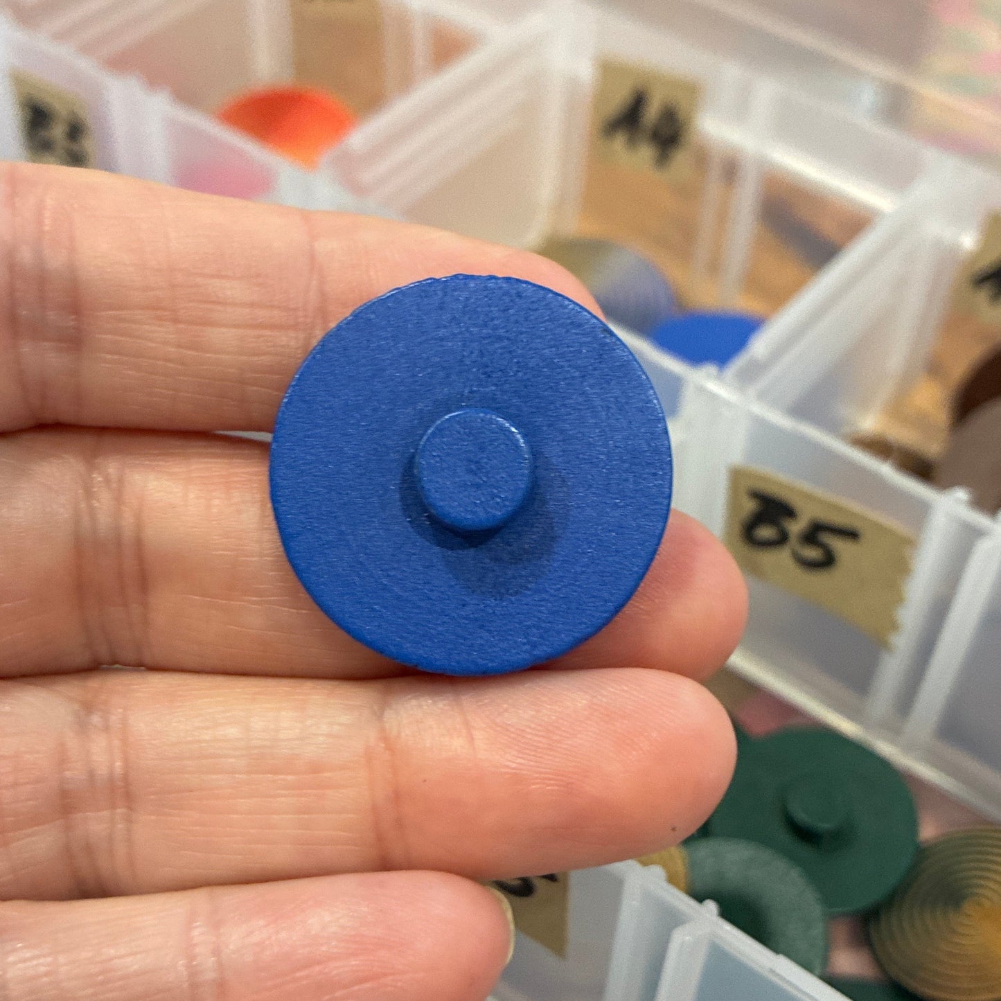 Hand holding a blue circular button with a transparent container of colorful round objects in the background.