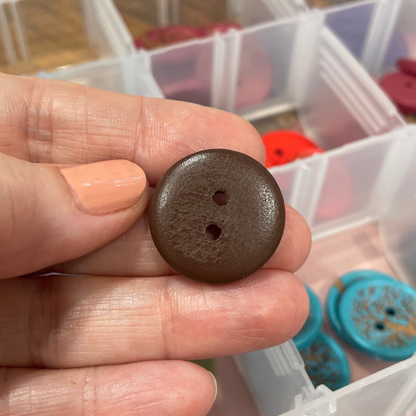 Hand holding a brown button with a clear plastic container of buttons in the background