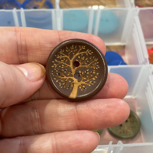Hand holding a button with a tree design in front of a container of assorted buttons.