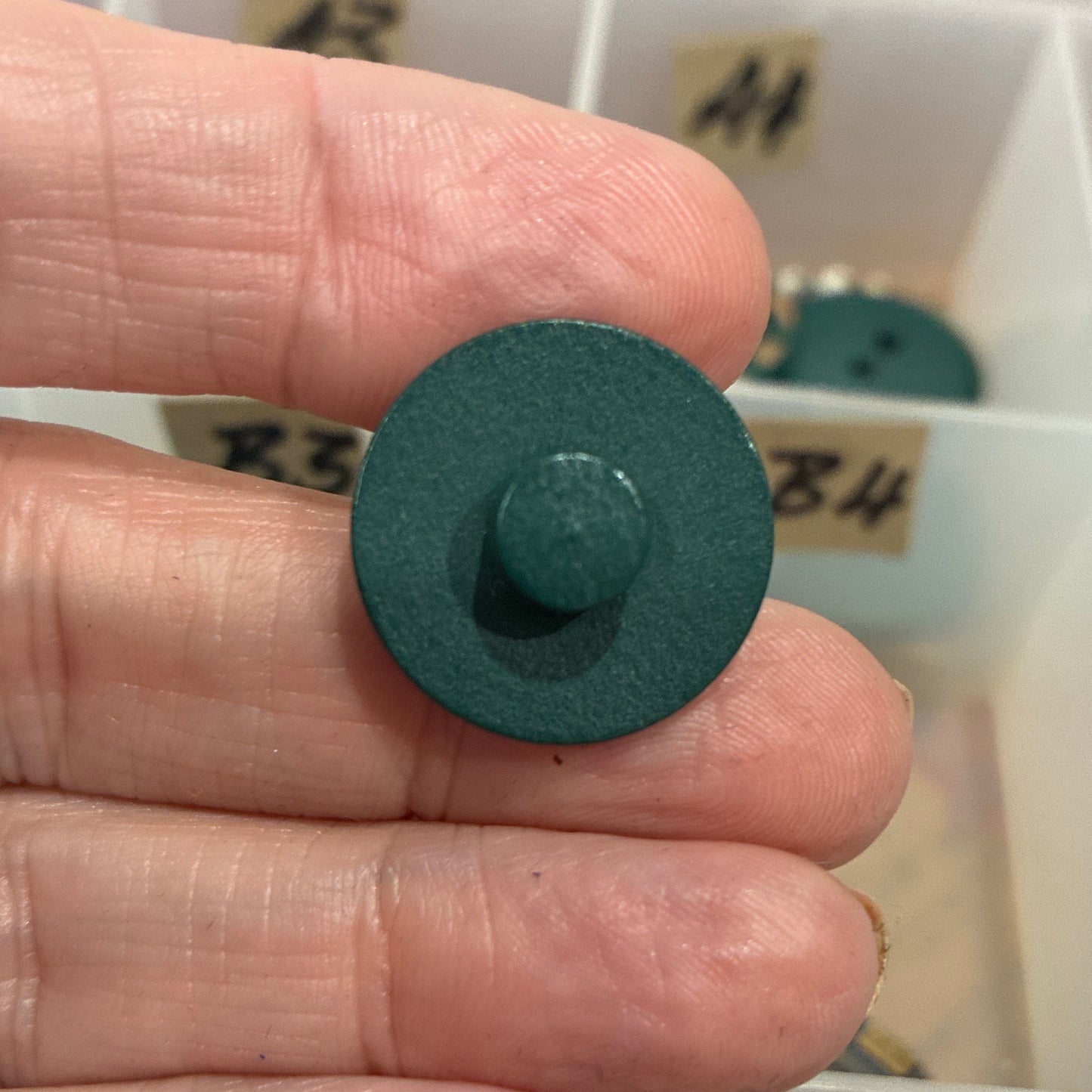 Green button held between fingers with a container of buttons in the background