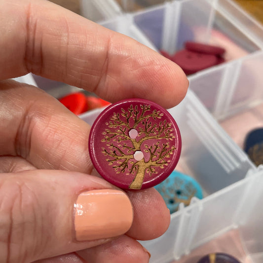 Hand holding a decorative button with intricate design, surrounded by a clear container with more buttons.