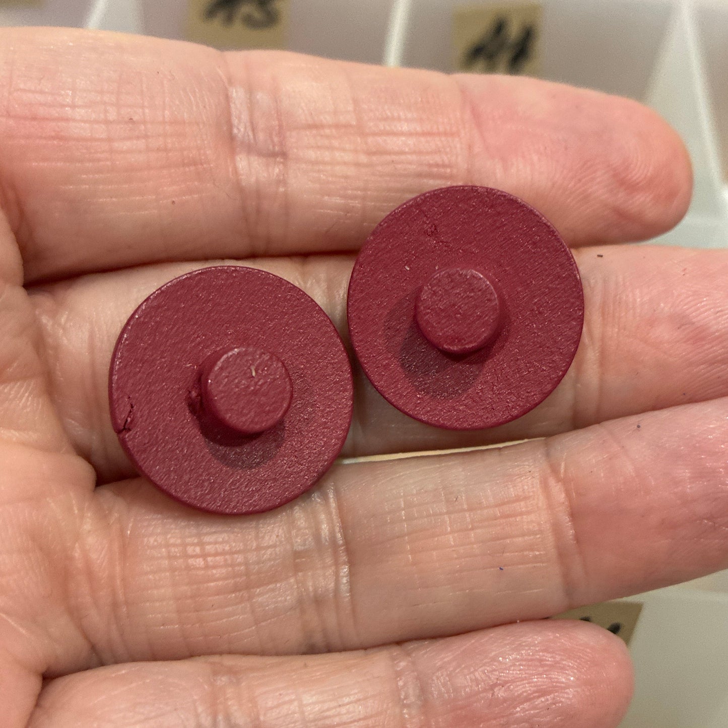 Two red circular objects held between fingers with a container of buttons in the background