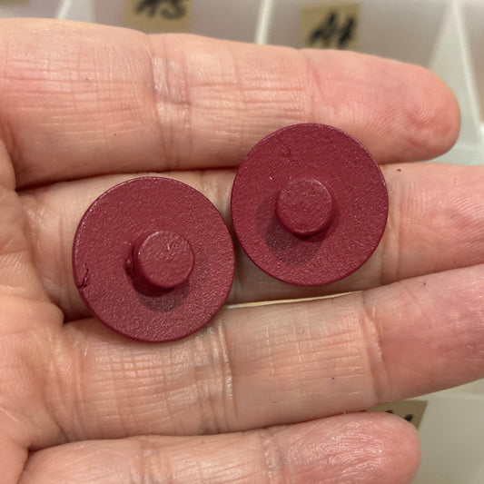 Two red circular objects held between fingers with a container of buttons in the background