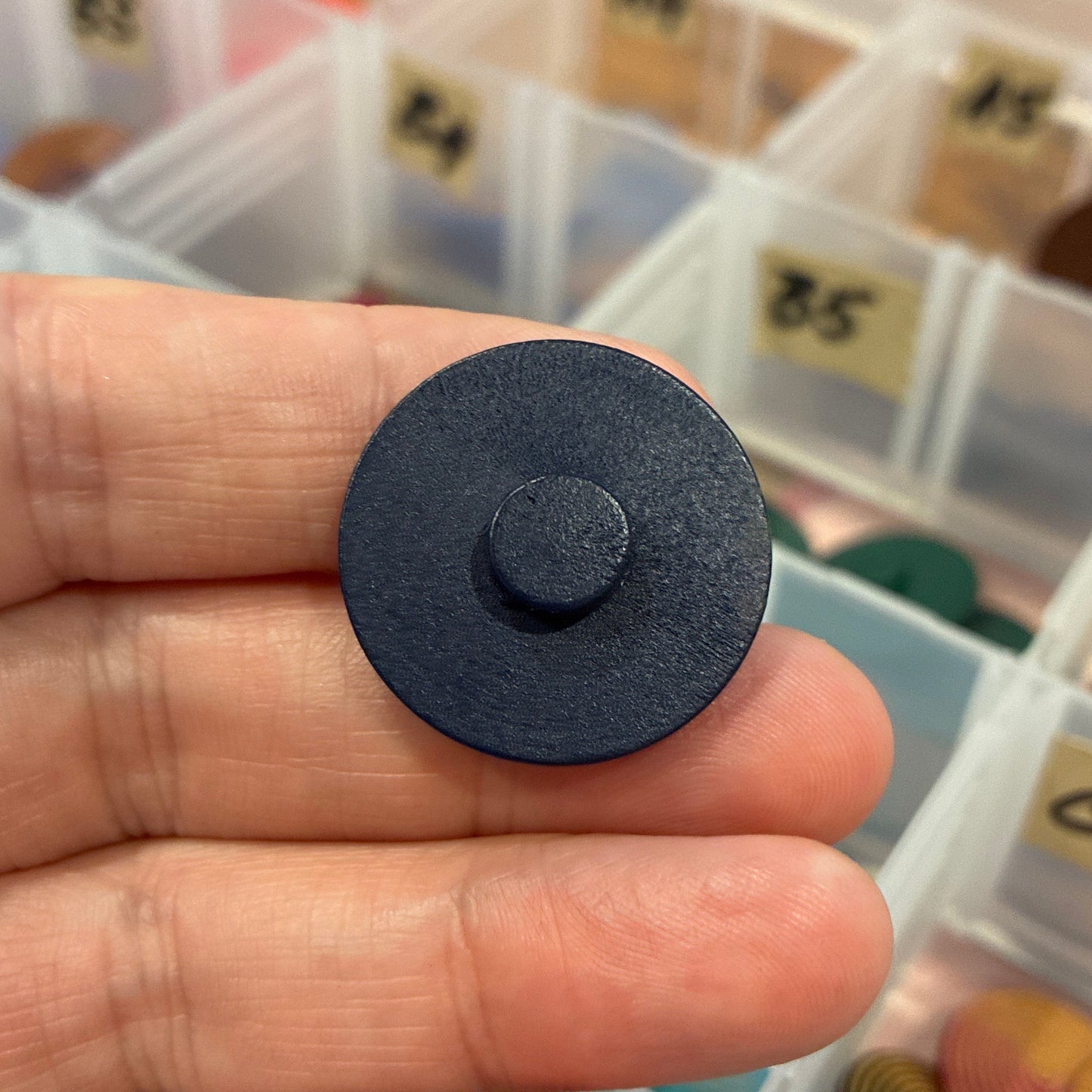 Black round button held in a hand with a container of assorted buttons in the background