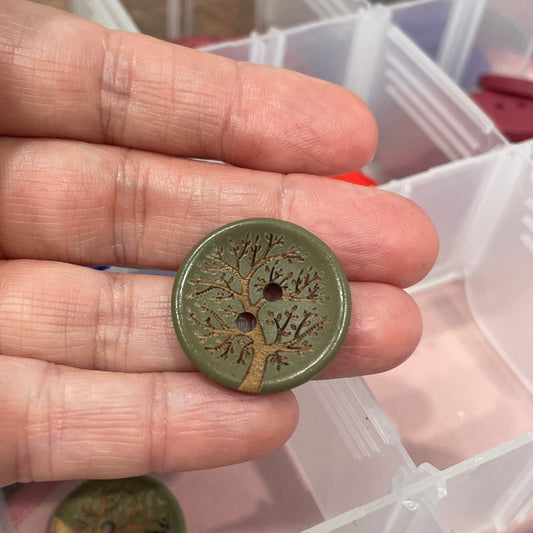 Hand holding a green button with a detailed design, against a background of a clear plastic container with more buttons.