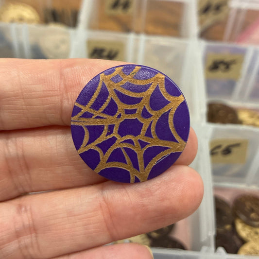 Hand holding a purple button with gold spider web design in front of a container of buttons.