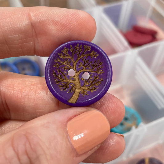Hand holding a purple button with a tree design, with a container of buttons in the background.