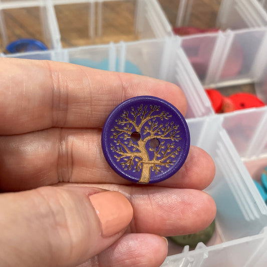 Hand holding a purple button with a tree design in front of a container of buttons.