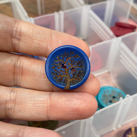 Hand holding a decorative button with intricate design, surrounded by various buttons in a clear container.