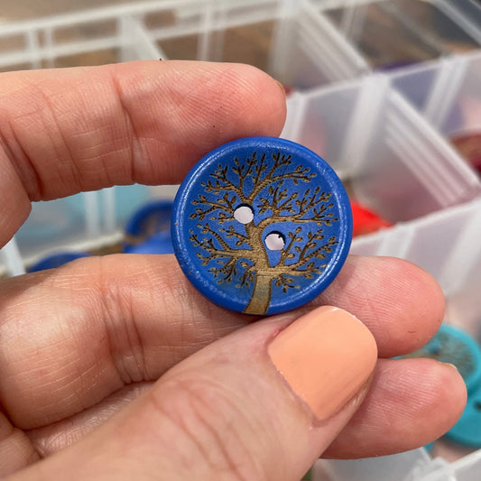Hand holding a blue button with a tree design in front of a container of buttons.