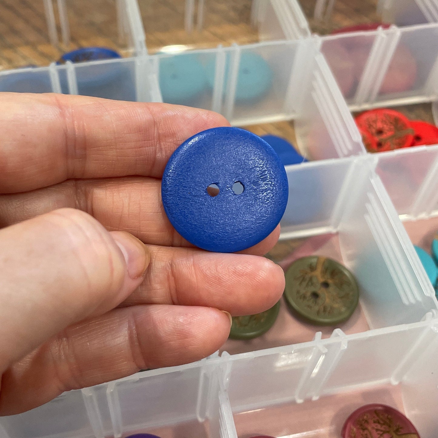 Hand holding a blue button with a background of assorted buttons in containers
