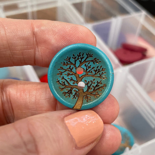 Hand holding a decorative button with intricate design, with a clear container of buttons in the background.